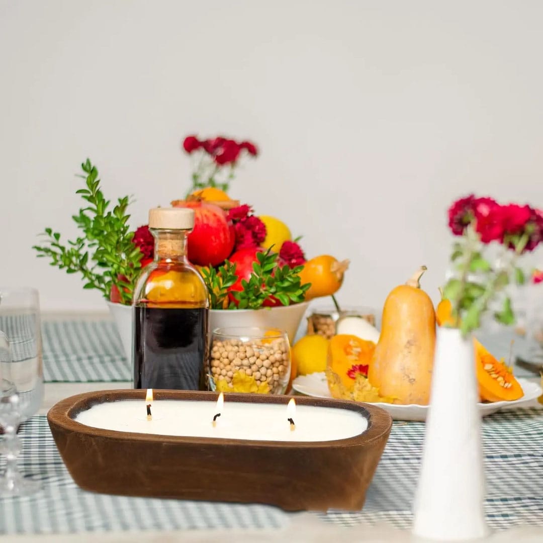 A table with a three-wick candle on wooden candle holders, a vase with flowers, a bottle with dark liquid, a bowl of chickpeas, squash, pomegranate, and other fruits creates the perfect restaurant ambiance.