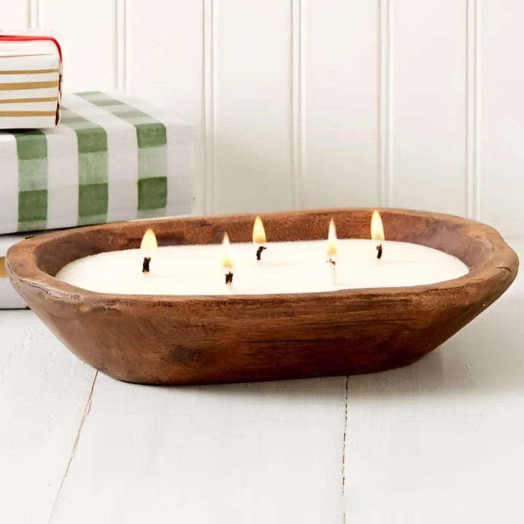 A wooden bowl filled with white wax and five lit candle wicks sits on a white surface near stacked books, adding the cozy charm of wooden candle holders to your space.
