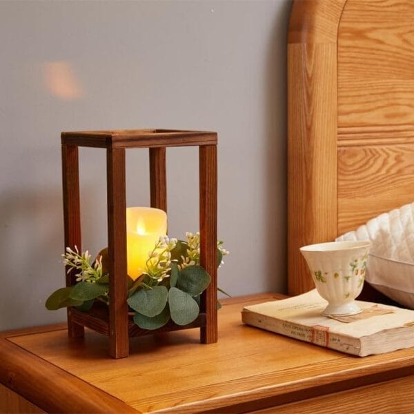 A wooden lantern-style candle holder, perfect as wood wedding table decor, sits on a bedside table with greenery, a teacup, saucer, and a closed book.