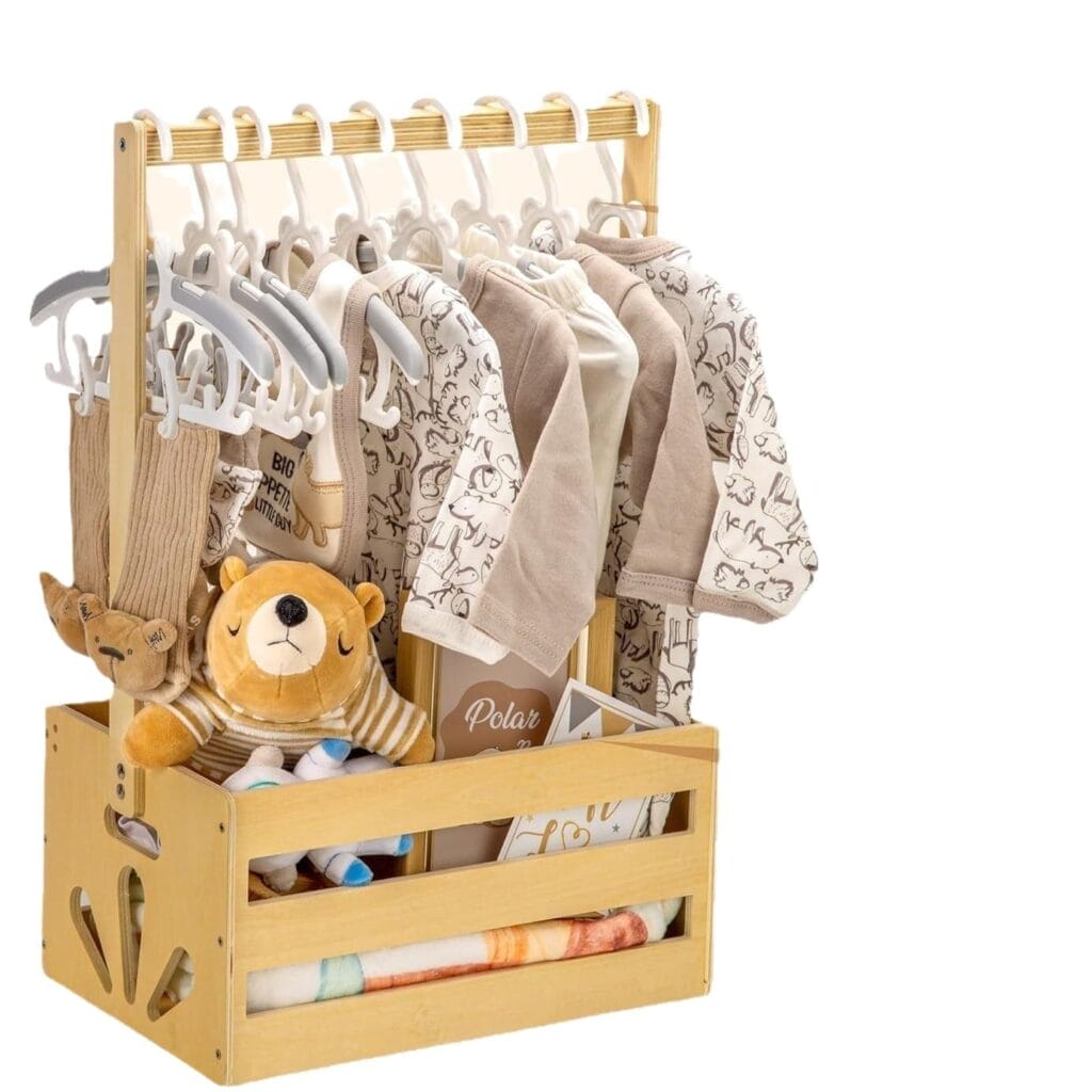 Wooden crate with baby clothes hanging on a small wooden coat holder, a plush toy, rolled blanket, and other baby items neatly arranged inside and around the crate.