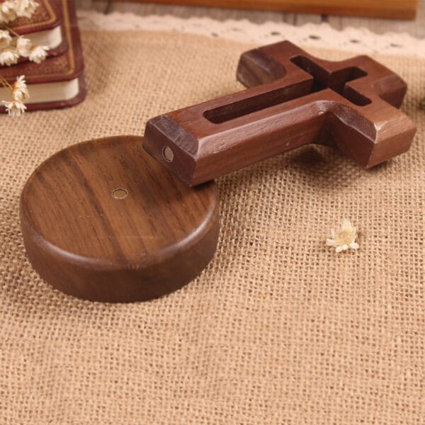 A wooden cross-shaped object with a round wooden base rests on a burlap surface, perfect as a DIY decorative craft, with dried flowers and a book in the background.