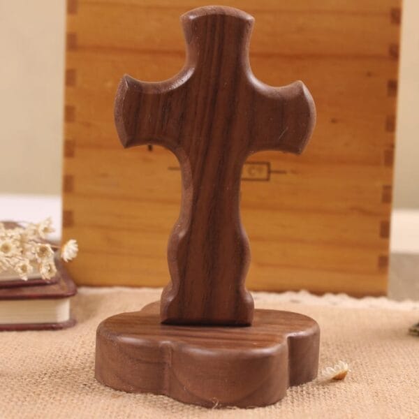 A wooden cross stands upright on a matching wooden base, perfect as a DIY decorative craft, placed on a textured surface with a book and flowers in the background. Available from global distributors.