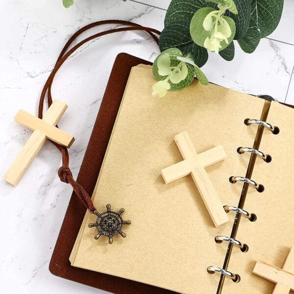 An open ring-bound notebook with wholesale wooden cross pendants, a leather cord featuring a laser cut metal charm, and green leaves on a marble surface.