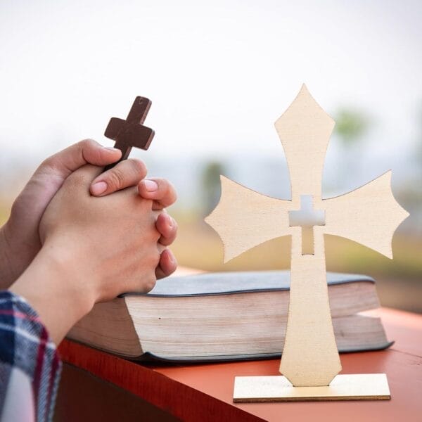 Two hands holding a small custom wooden cross above a closed Bible, with a larger wooden cross decoration standing upright on a table in the foreground—a perfect touch for meaningful home decor.