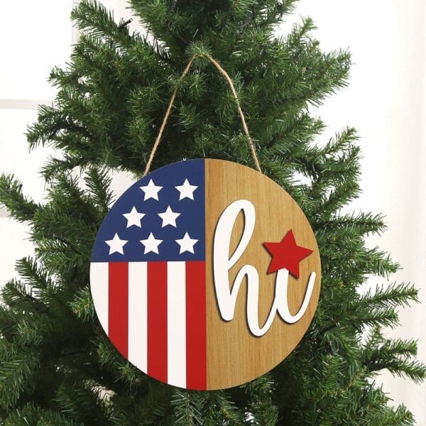 A round wooden Independence Day hanging sign with the American flag on the left and "hi" with a red star on the right, displayed on a green Christmas tree—perfect custom wood decor from a wholesale manufacturer.