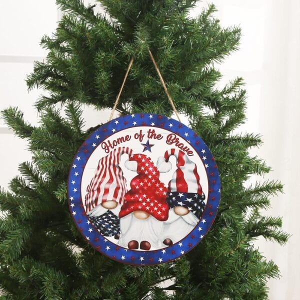 A round Independence Day hanging sign with three gnomes in patriotic hats and the text "Home of the Brave" decorates a Christmas tree. The blue border with white stars highlights this custom wood decor, perfect from any wholesale manufacturer.