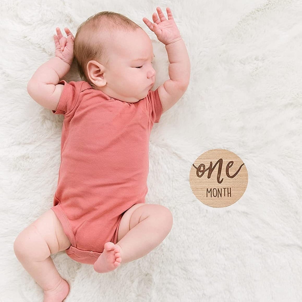A baby in a pink onesie lies on a white furry blanket with a custom wooden tag next to them that reads "one month.