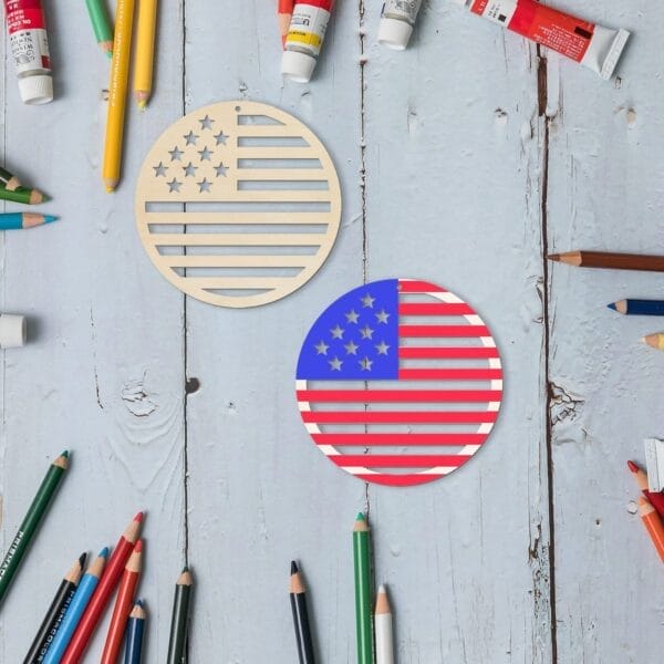 Two round cutouts of the American flag, perfect for American Independence Day decoration, rest on a wooden surface surrounded by colored pencils and paint tubes, showcasing creativity from a leading wooden flags manufacturer.