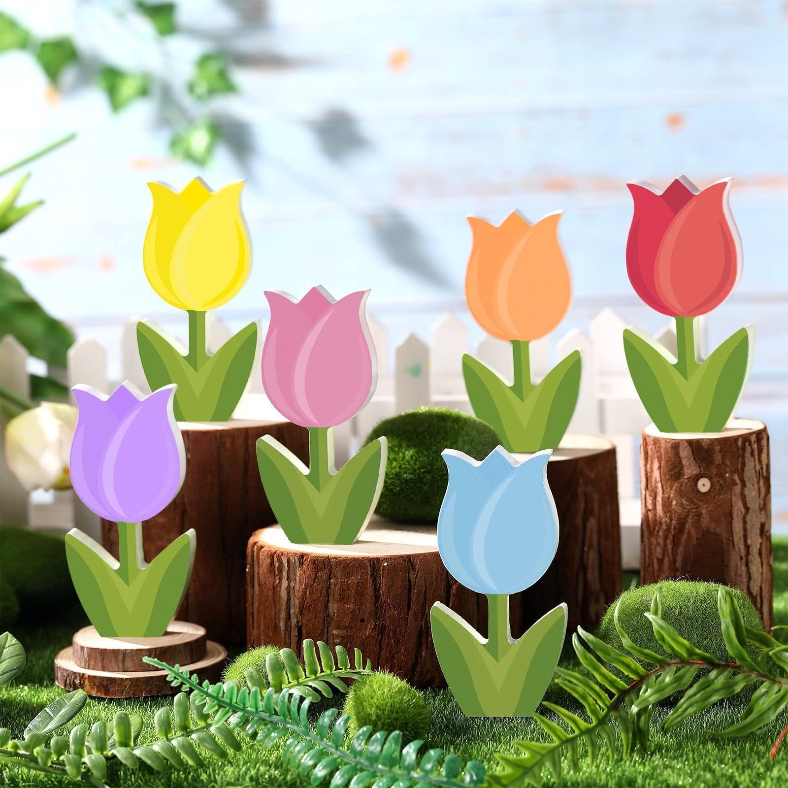 Six colorful wooden flowers shaped like tulips are displayed on tree stumps and moss, with a white picket fence and blurred outdoor background.