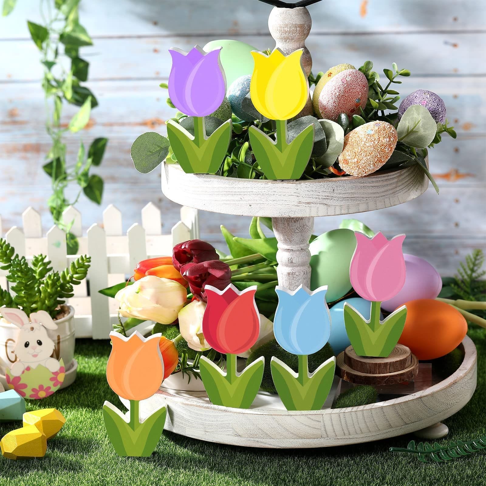 A two-tiered wooden tray holds colorful Easter eggs, artificial tulips, and wooden flowers, set on grass with a picket fence and a bunny decoration in the background—a perfect Layered Tray Craft for Mother's Day.