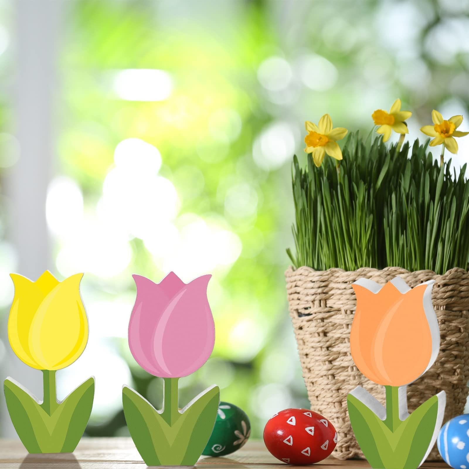 Three colorful tulip decorations and painted Easter eggs are arranged next to a wicker basket of grass with blooming yellow daffodils, creating a charming Mother's Day scene featuring wooden flowers.