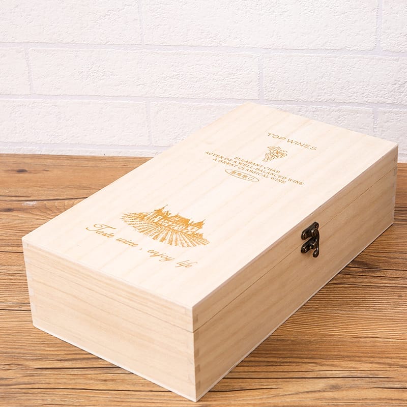 A rectangular custom wooden wine box with a hinged lid, engraved text, and a clasp, placed on a wooden surface against a white brick wall.