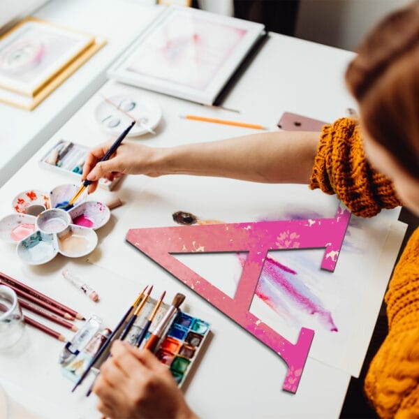 A person painting a large pink wooden craft letter "A" on paper, surrounded by watercolor paints, brushes, and art supplies on a white table—perfect for DIY wall decor projects.