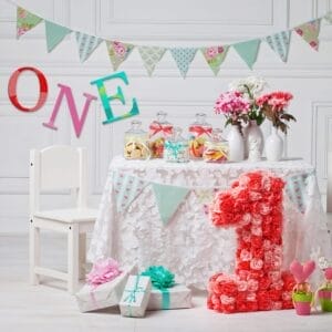 A decorated table with flowers, candy jars, wrapped gifts, a large pink number 1, DIY wall decor, and Wooden craft letters spelling "ONE," suggesting a first birthday celebration.