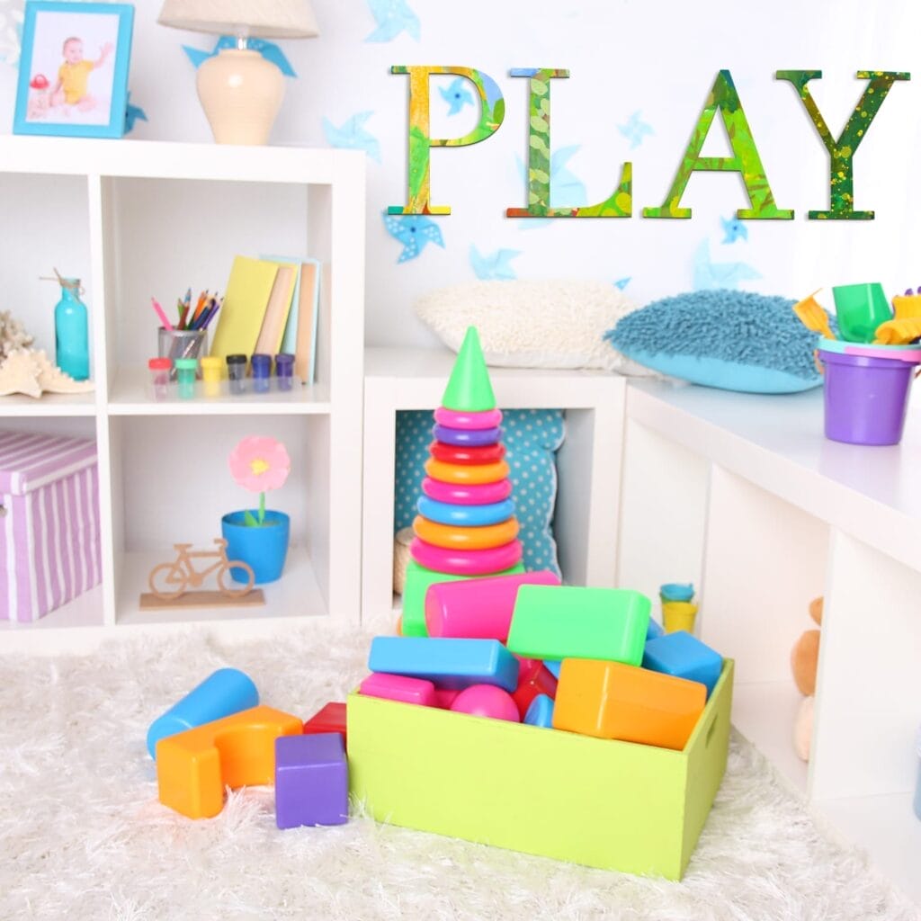 A brightly colored playroom with a green box of plastic blocks, a stacking ring toy, shelves with toys, and DIY wooden craft letters spelling "PLAY" as wall decor.