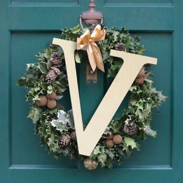 A green wreath with pinecones, a gold bow, and a large beige wooden craft letter "V" hangs on a dark green door—a charming piece of wall decor perfect for wholesale or home display.