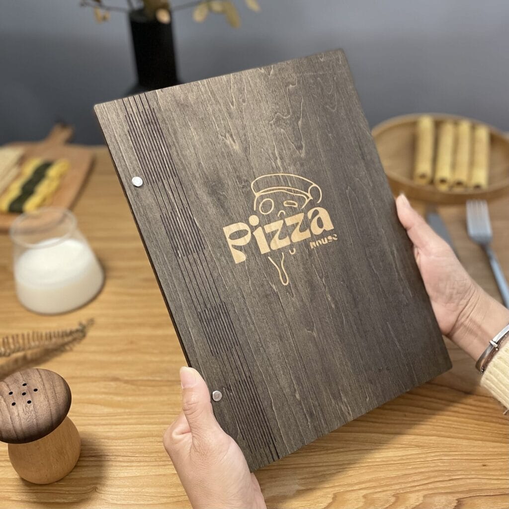 A person holds a wooden menu with "Pizza" engraved on the cover at a restaurant table set with a drink, rolled food, fork, pepper shaker, and decorative plant. The elegant wooden menu adds rustic charm to the inviting dining scene.