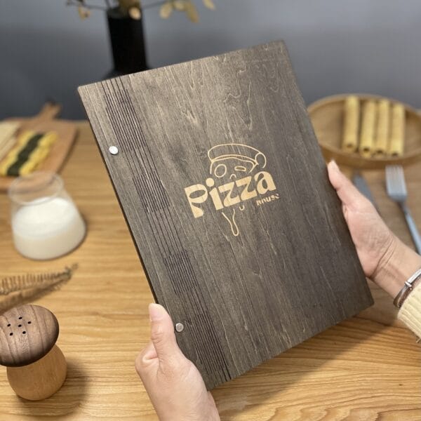 A person holds a wooden menu with "Pizza" engraved on the cover at a restaurant table set with a drink, rolled food, fork, pepper shaker, and decorative plant. The elegant wooden menu adds rustic charm to the inviting dining scene.