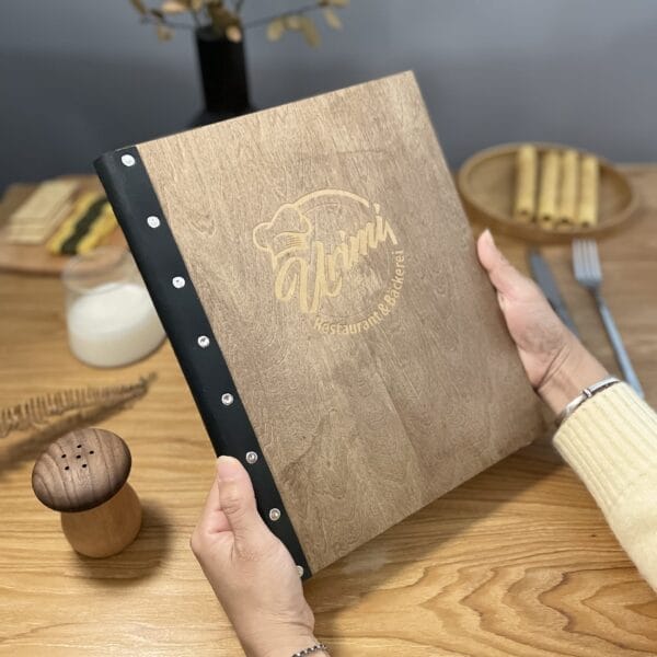 Two hands hold an eco-friendly, wooden menu with “Yumi Restaurant & Bäckerei” engraved on the cover, resting on a wooden table set with cutlery, a salt shaker, and dishes.