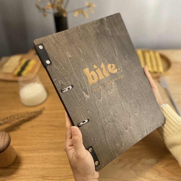A person holds an eco-friendly wooden menu engraved with "bite food & beverage" at a table with plates of food and a glass of milk in the background.