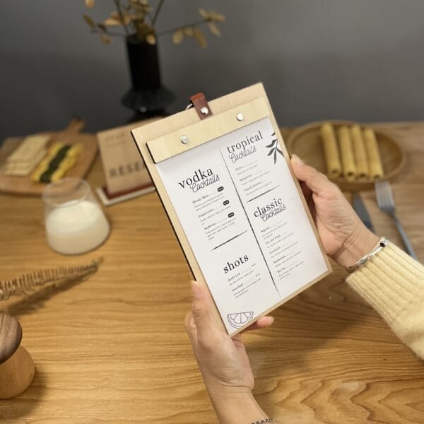 Person holding a wooden menu board with drink options: Vodka, Tropical, Classic cocktails, and Shots. Eco-friendly design.