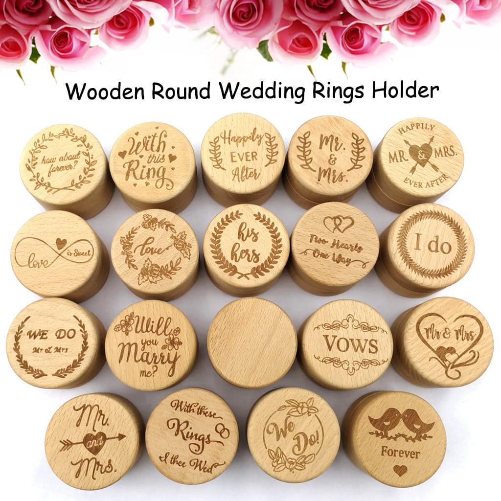 A set of 18 round wooden ring holders and wooden ring box designs with engraved phrases, arranged in rows on a white surface with pink roses at the top.
