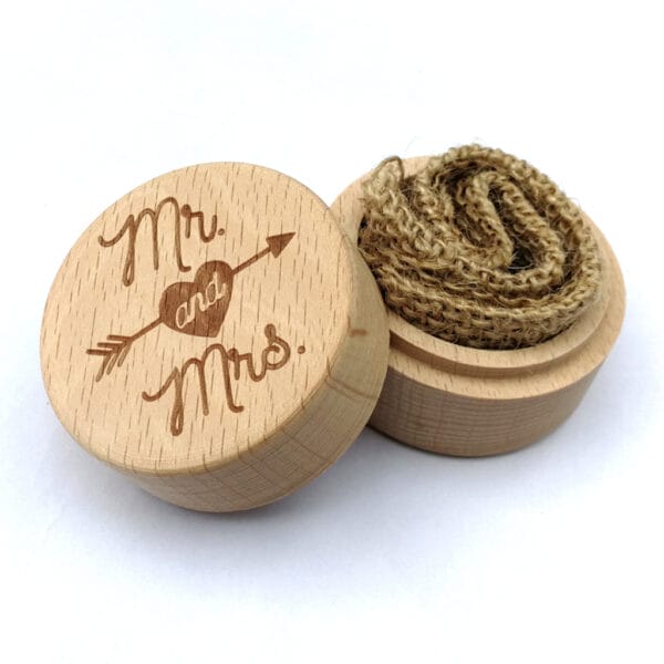 A round wooden ring box with "Mr. and Mrs." and an arrow engraved on the lid, shown open with a piece of burlap fabric inside—perfect engraved packaging for wedding rings.