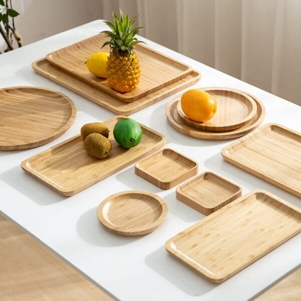 Assorted rectangular and round bamboo trays and a wooden serving tray are displayed on a white table, with a pineapple, lemons, kiwis, and a lime placed on some of the trays.