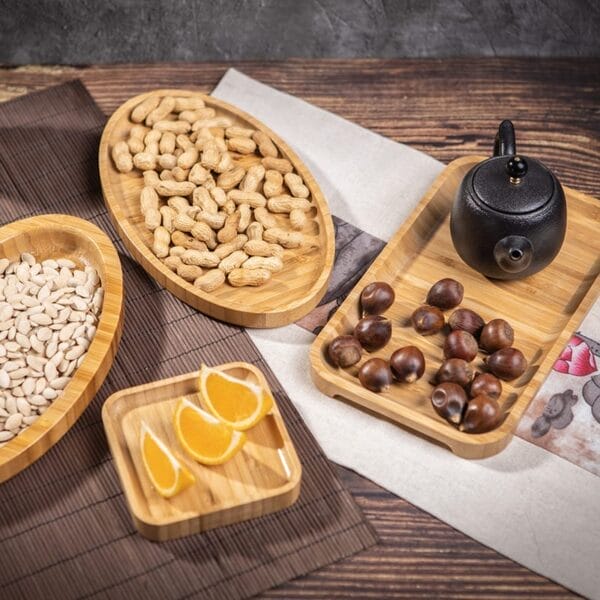 Assorted bamboo snack trays with peanuts, chestnuts, orange slices, and a black teapot are arranged on a wooden table with placemats.