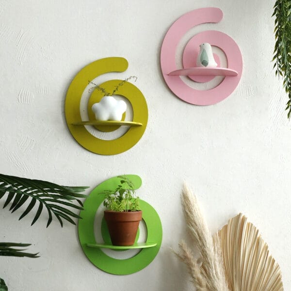Three spiral-shaped wooden stands in yellow, pink, and green hold small decorative objects and a potted plant, mounted on a white wall with surrounding plants.