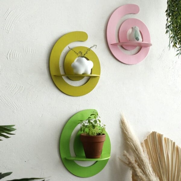Three circular Wall Crystal Storage Shelves in yellow, pink, and green hold small decorative objects and a potted plant against a white textured wall, with lush plants in the foreground. Perfect for stylish organization from a wholesale supplier.