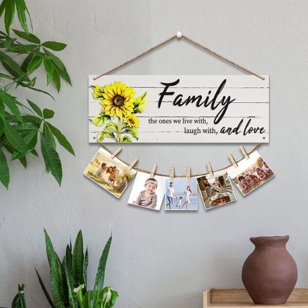 A wooden wall decor display features a "Family" sign with a sunflower, five family photos clipped below, and houseplants nearby on a shelf and the floor.