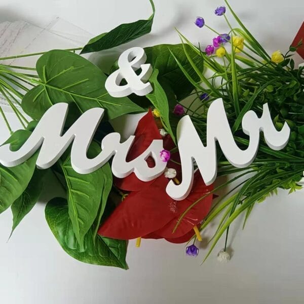 White "Mr & Mrs" wooden wedding decorations rest on green and red artificial leaves, accented by small purple and white flowers, all set against a crisp white background.