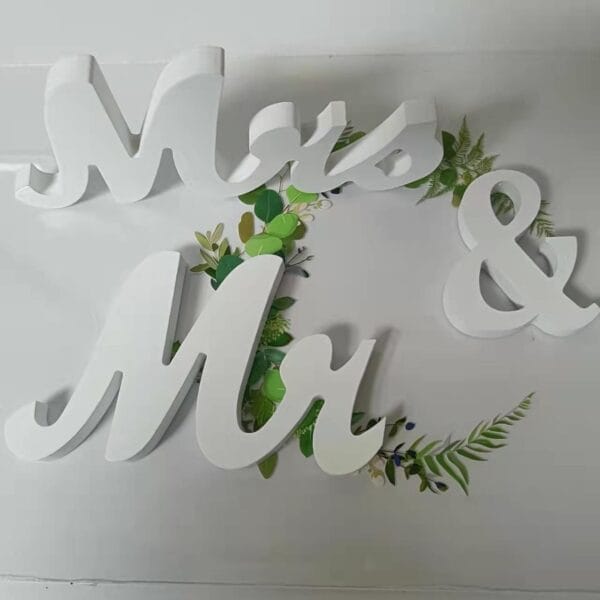 White 3D letters spelling "Mrs & Mr" are placed on a flat surface with green leaves and ferns underneath, making charming wedding signage and party signage props.
