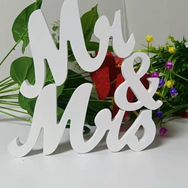 White "Mr & Mrs" party signage in cursive font stands in front of a bouquet of green leaves and colorful flowers on a white surface, making it a charming choice for wooden wedding decorations.