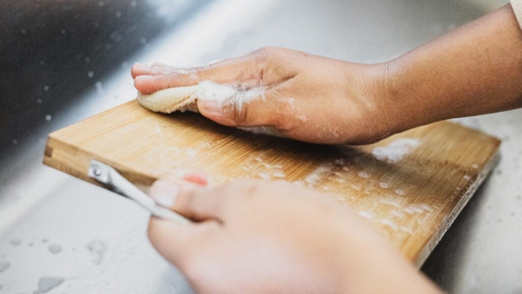 Hands washing a bamboo cutting board with soapy sponge. Safe home sanitization.
