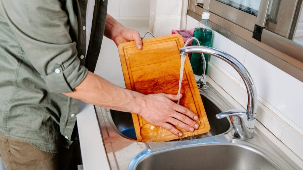 Person washing a wooden cutting board under running water, demonstrating how to sanitize a wooden cutting board safely at home.