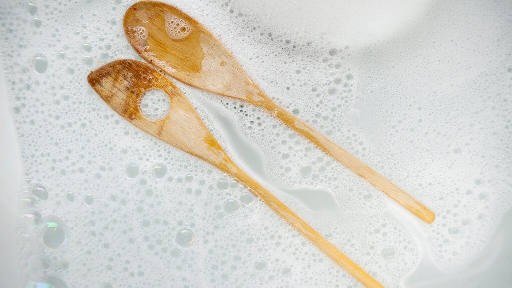 Two wooden spoons soaking in soapy water; How to Sanitize Wooden Spoons