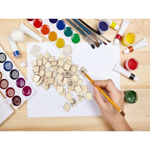 Person painting floral designs on blank square wood cutouts. Craft supplies surround them.
