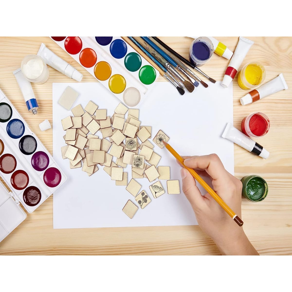 Person painting floral designs on blank square wood cutouts. Craft supplies surround them.