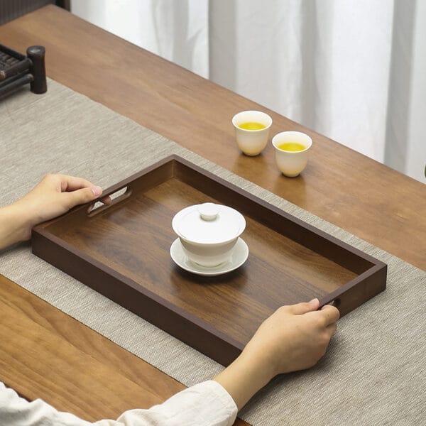 Person holding a dark brown wooden serving tray with teacups and a lidded bowl. Eco-friendly wooden tray.