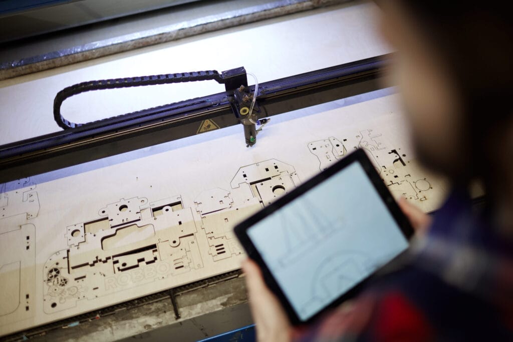 Man using a tablet to oversee a laser cutter working on a wood project. "ROBBO" visible on material.
