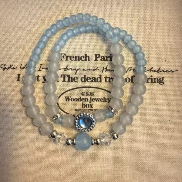 Two light blue beaded bracelets, one with decorative centerpiece and silver accents, on textured fabric.