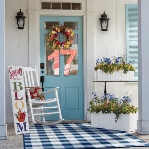 Charming spring porch decor: blue door, floral wreath, rocking chair, and tiered flower planters.