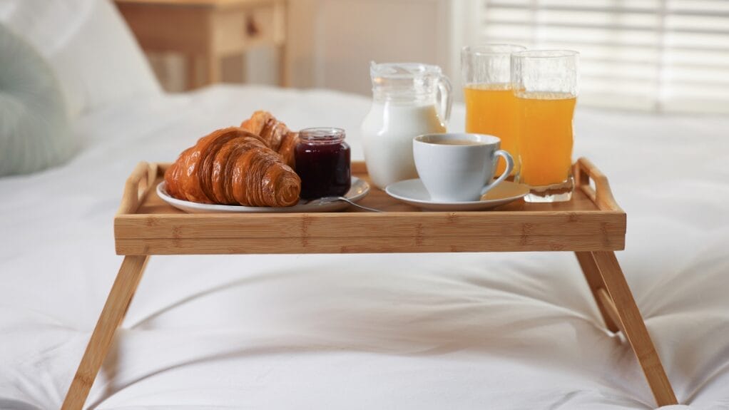 Breakfast on serving tray: croissants, jam, coffee, milk, and orange juice on a bed.