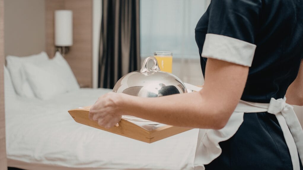 Hotel maid carrying a serving tray with a covered dish and juice in a hotel room.