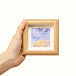 Hand holding mini square wooden frame showcasing an impressionist landscape. Wholesale wood picture frames available.