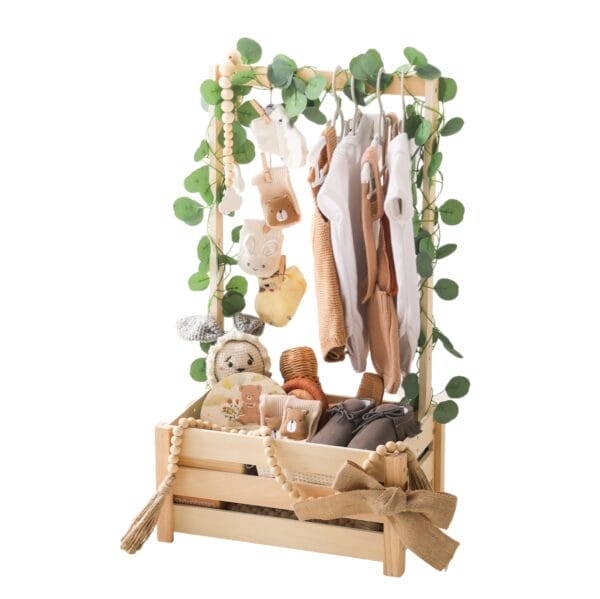 Baby clothes rack with neutral-toned outfits, shoes, and accessories in a wooden crate, decorated with greenery and beads.