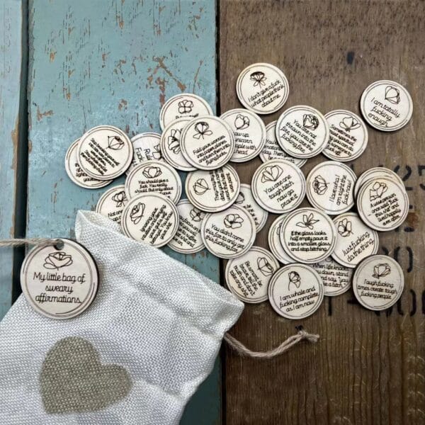 Wooden sweary affirmation tokens spilling from a drawstring bag with a heart design.