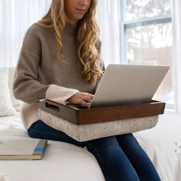 Woman uses laptop on a lap desk while sitting on a bed.